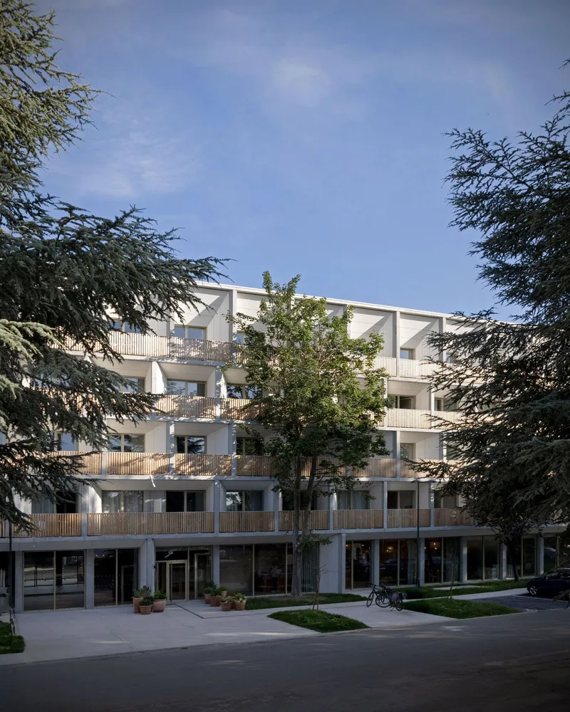 Courtyard facade with timber balcony railings and white concrete framed by mature trees under blue sky