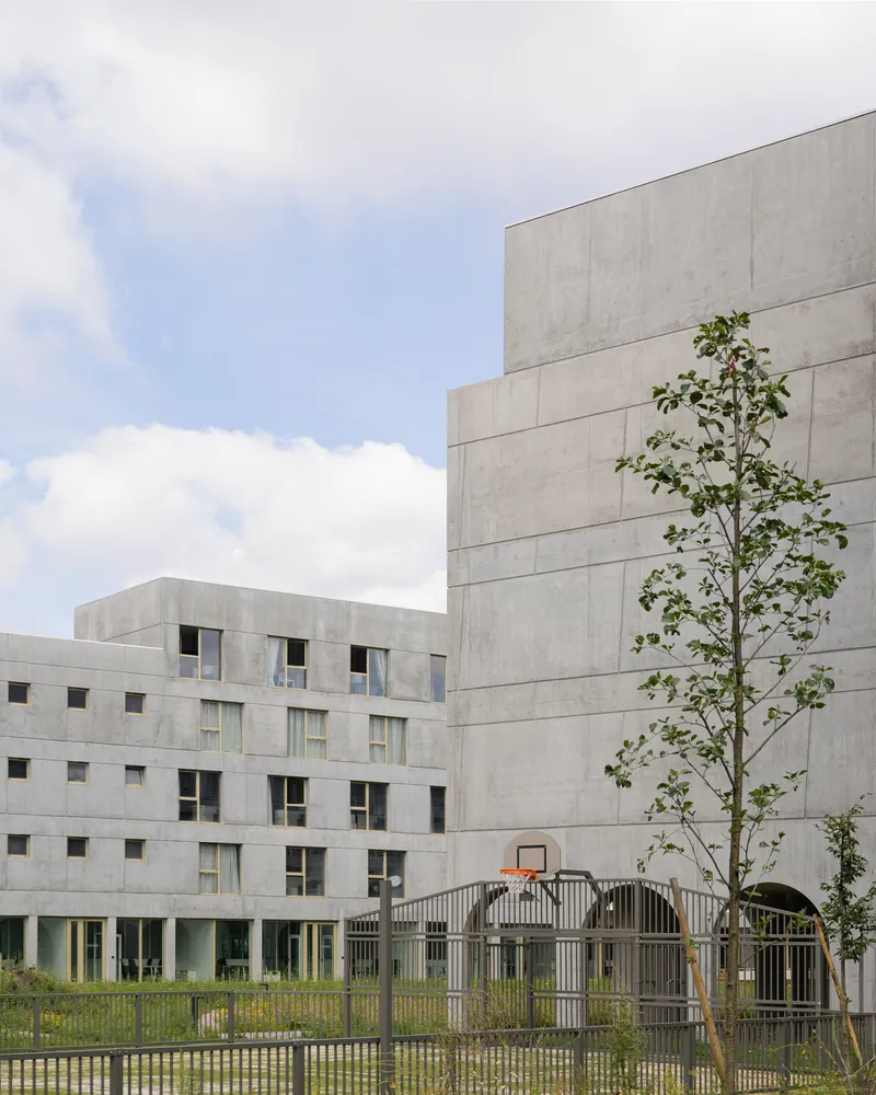 Concrete block facade with small punched windows beside a young tree and metal fenced play area