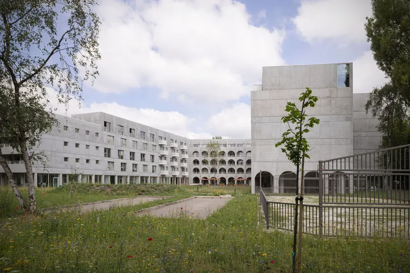 View across wildflower meadow toward concrete housing blocks with arched arcade and young tree
