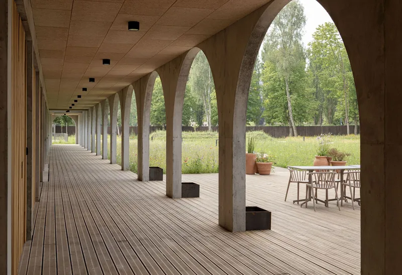 Covered timber deck walkway with concrete arches opening to wildflower meadow and potted plants