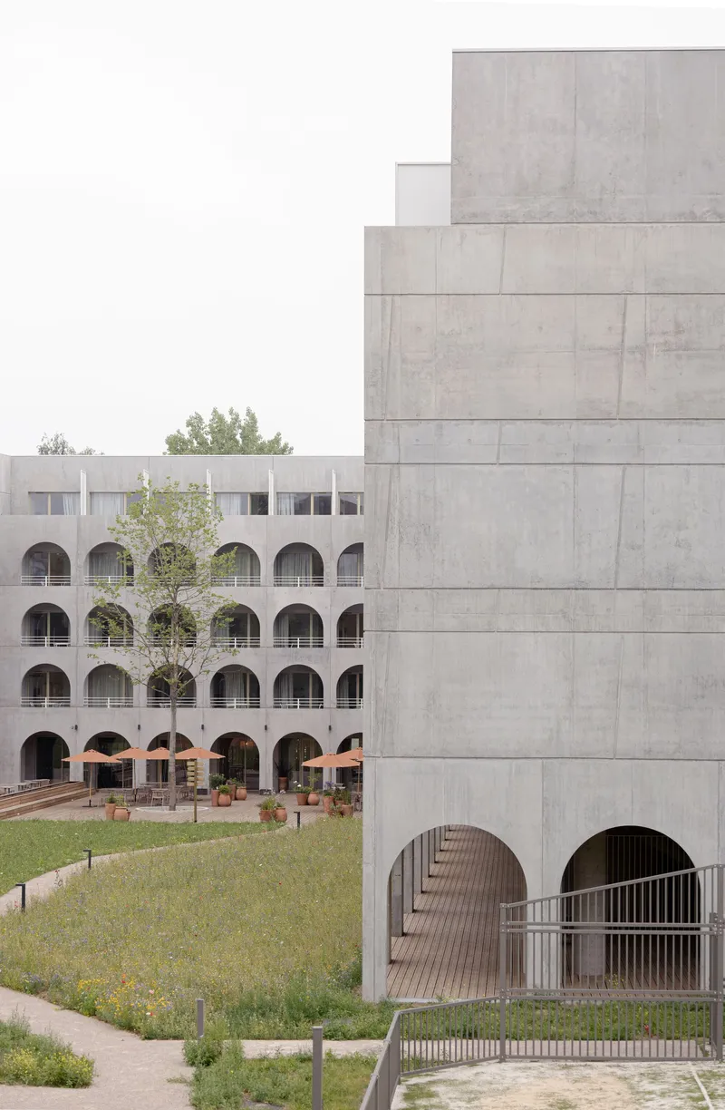 Concrete tower and arcade facades framing a wildflower meadow with a young tree in summer