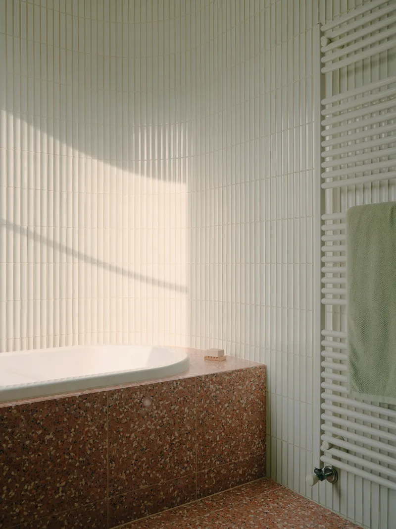 Bathtub alcove with terrazzo cladding and vertical ceramic tile catching afternoon sunlight
