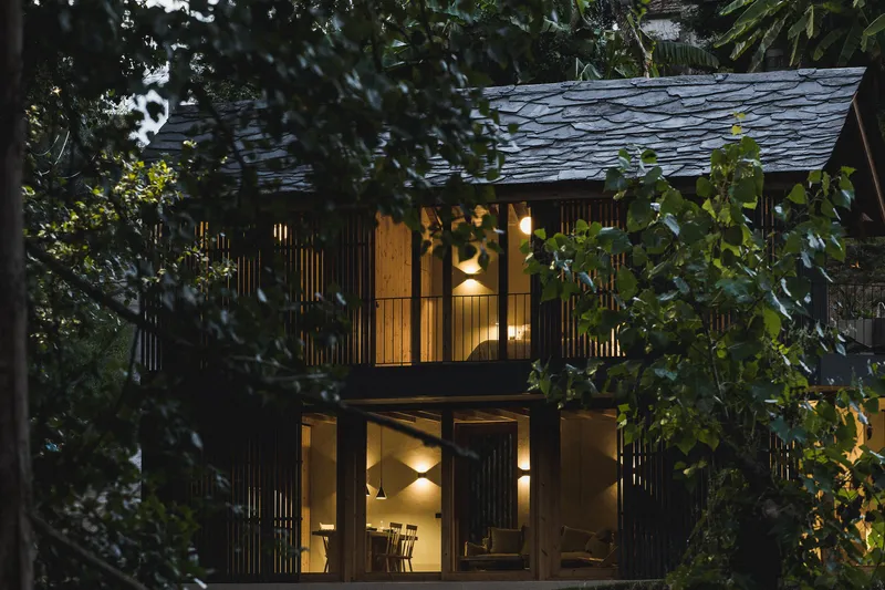Two-storey facade with vertical timber slats and slate roof at dusk surrounded by trees