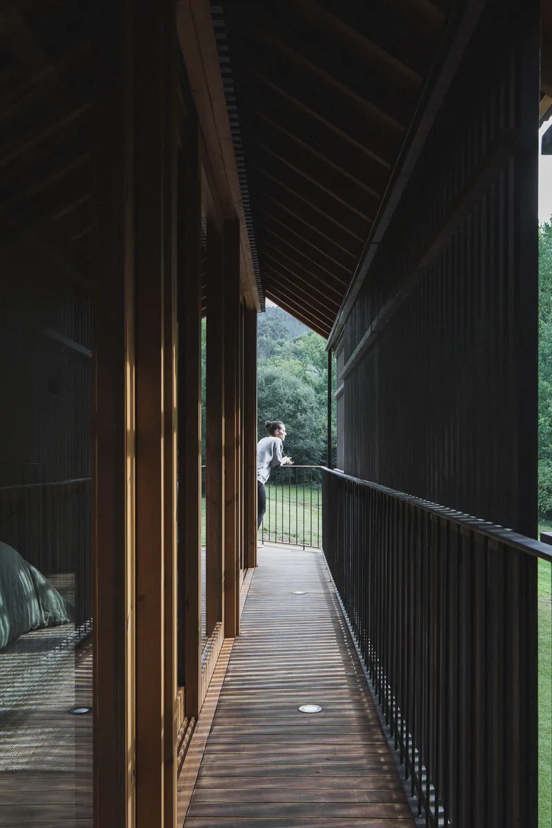 Narrow timber walkway with vertical slat walls leading to figure standing at exterior railing overlooking lawn