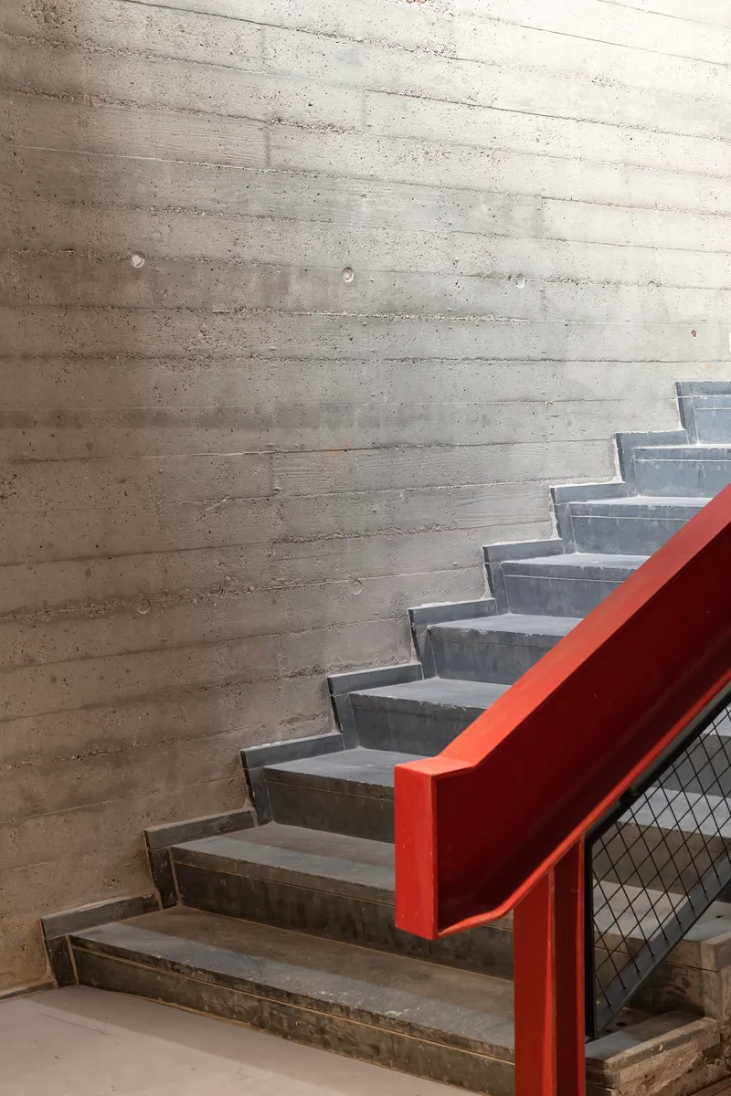 Concrete stair with red painted steel handrail ascending against a board-formed concrete wall