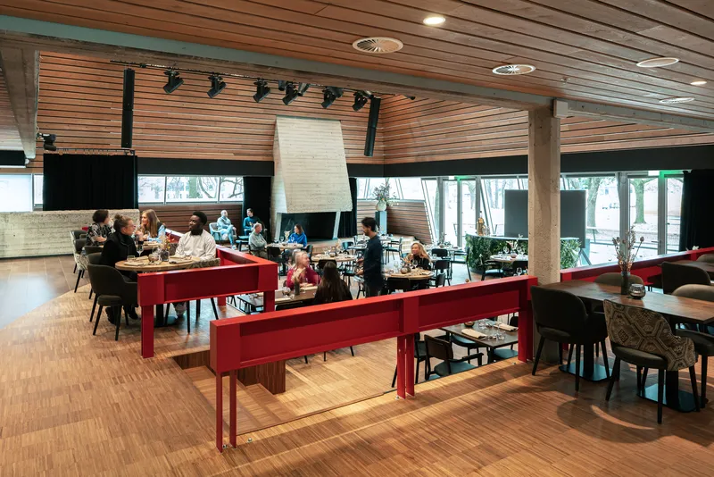 Communal dining space with red timber tables beneath slatted wood ceiling and concrete columns