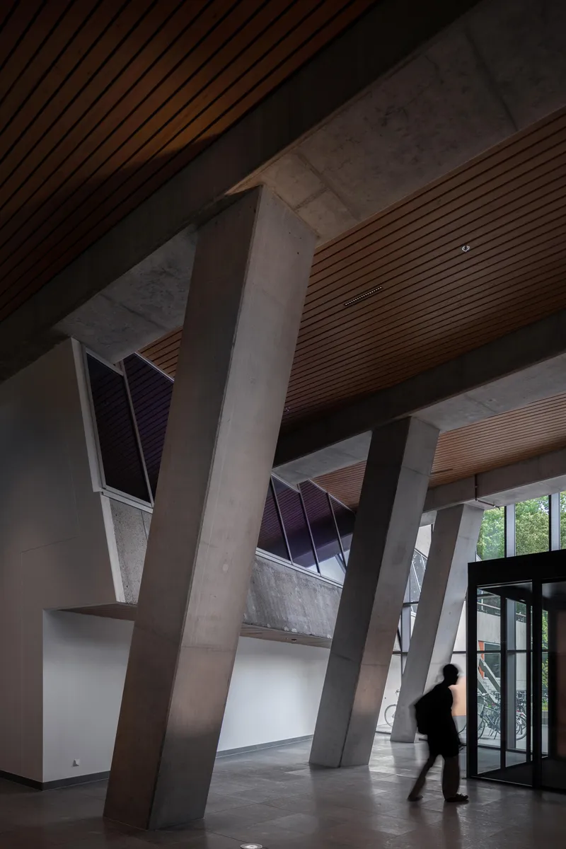Lobby interior with angled concrete columns and timber slat ceiling panels above figures passing through