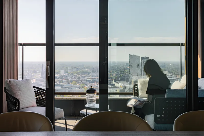 Interior view through floor-to-ceiling glazing with a figure reading on the balcony overlooking the cityscape