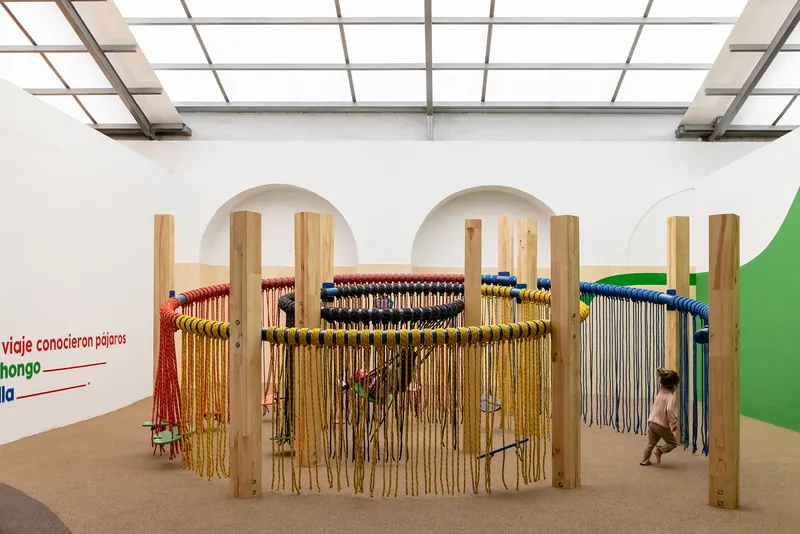 Timber-framed play structure with colored rope curtains beneath a gridded skylight as child runs through