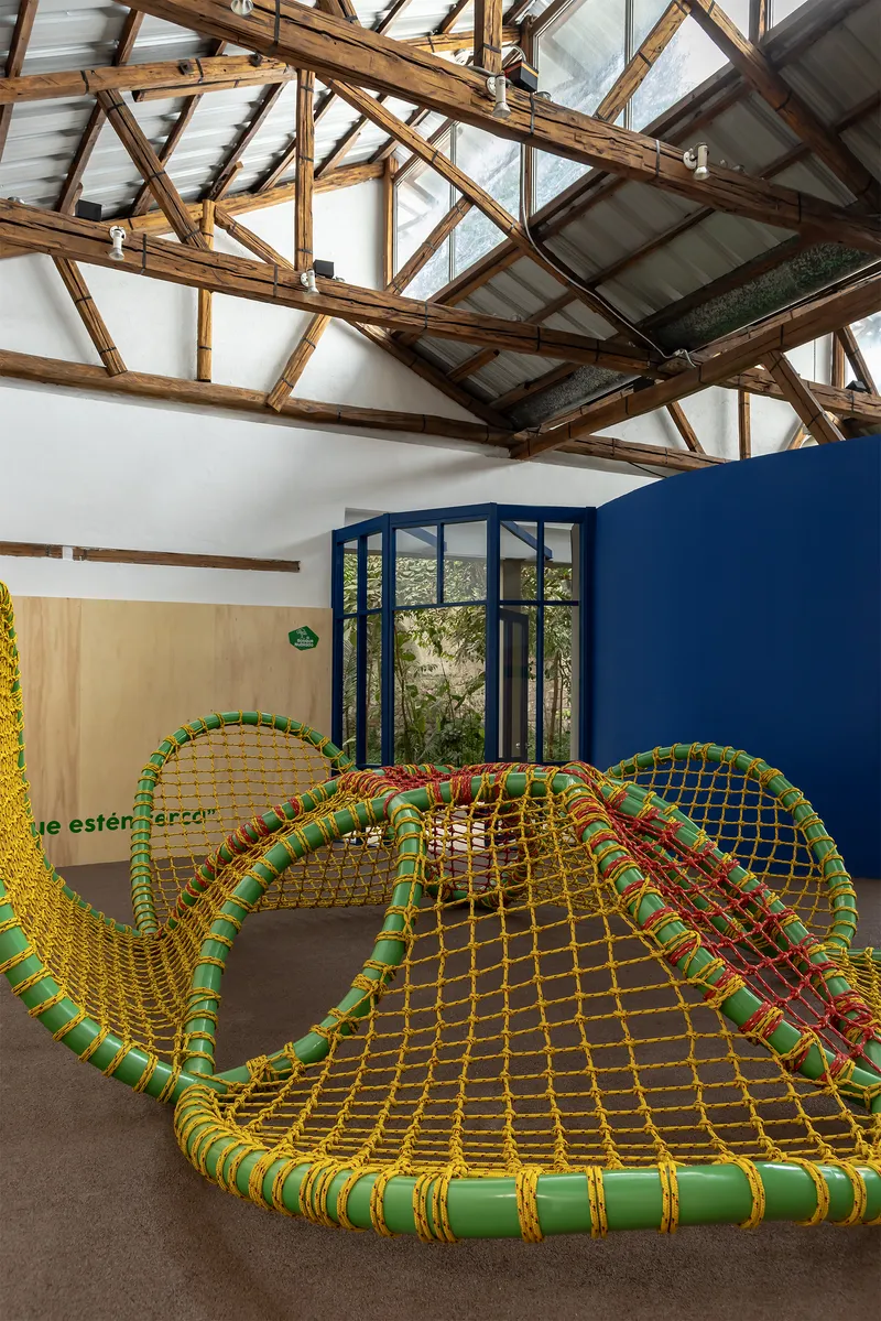 Woven rope climbing net in green and yellow beneath timber trusses and tall glazed doors