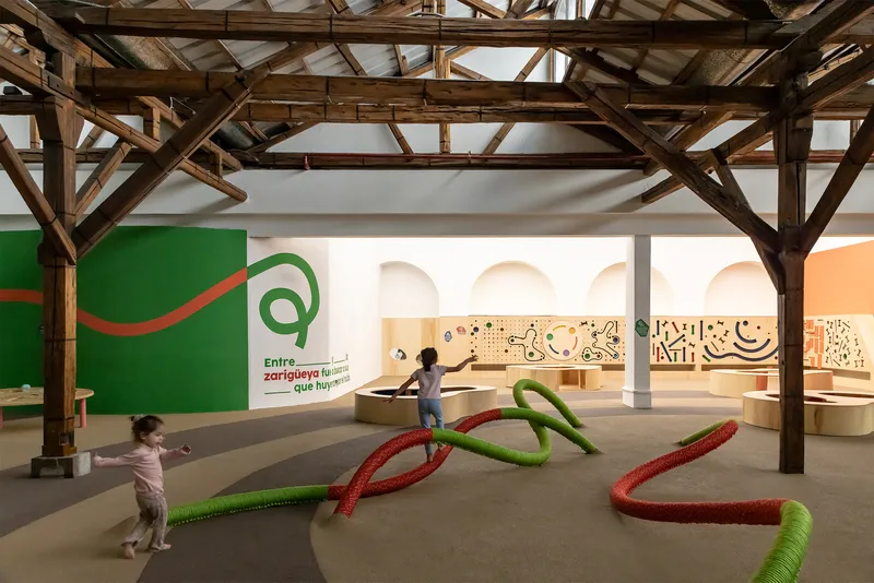 Children interacting with rope sculptures beneath timber trusses with wall graphics and circular niches beyond