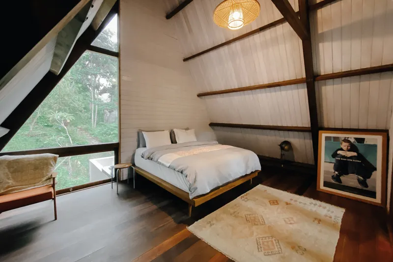 A-frame bedroom with exposed dark timber rafters and triangular glazing overlooking dense green forest