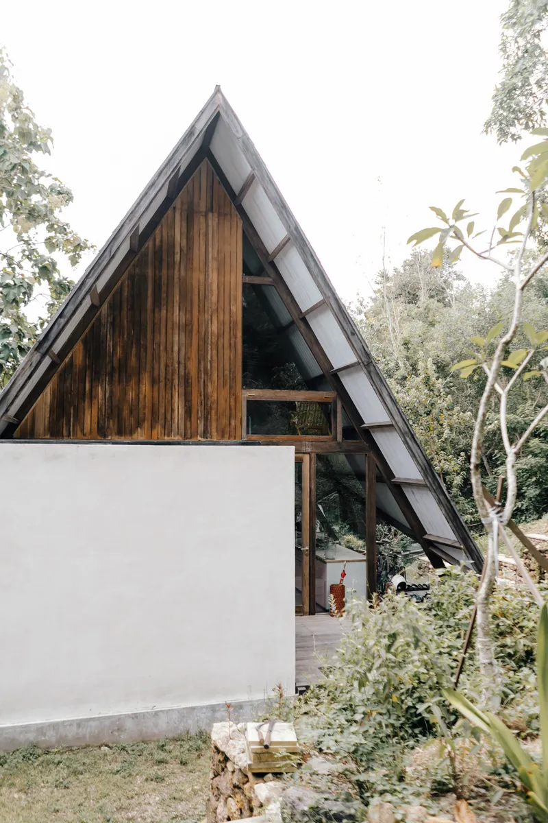 A-frame volume with dark timber gable and white masonry base wall seen through garden planting