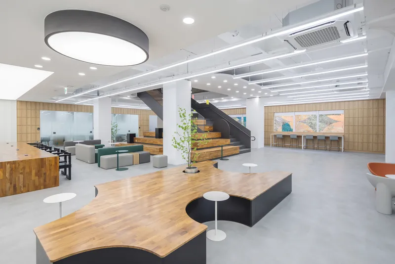 Open office interior with timber benches, black steel staircase and white columns under linear ceiling lights