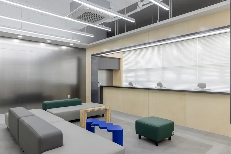 Waiting area with light wood service counter, translucent roller blinds and modular grey and green ottomans