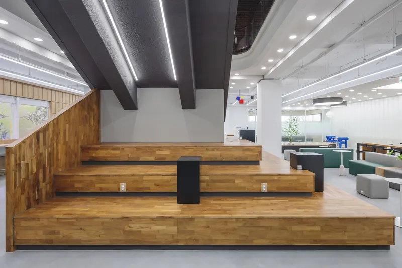 Tiered timber seating platform with steel beams overhead and exposed ceiling ducts in the office interior