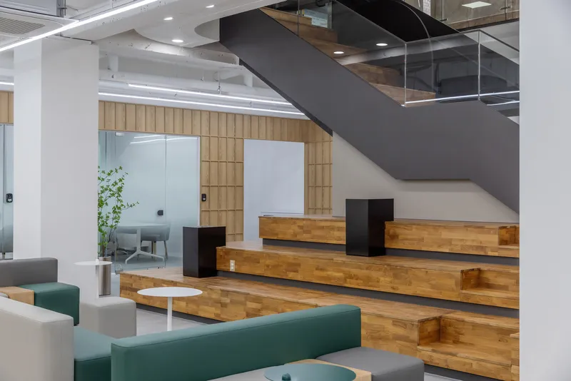 Open office lounge with timber stepped seating, black steel stair, and green upholstered benches beside potted greenery