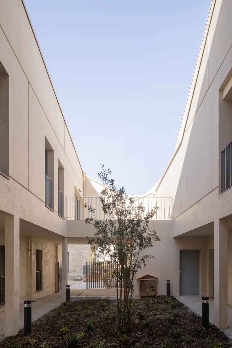 Narrow courtyard passage between two-story concrete facades with metal railings and a young tree in a planted bed