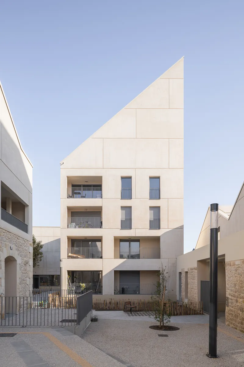 Angled facade with diagonal roofline clad in pale stone panels and recessed balconies facing a gravel courtyard