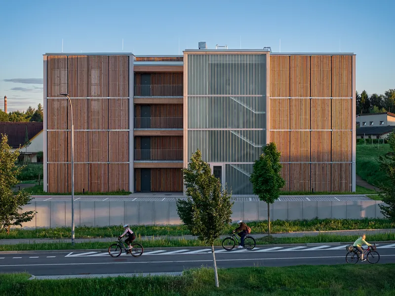 Street facade with alternating timber and translucent panel screens as three cyclists pass at dusk