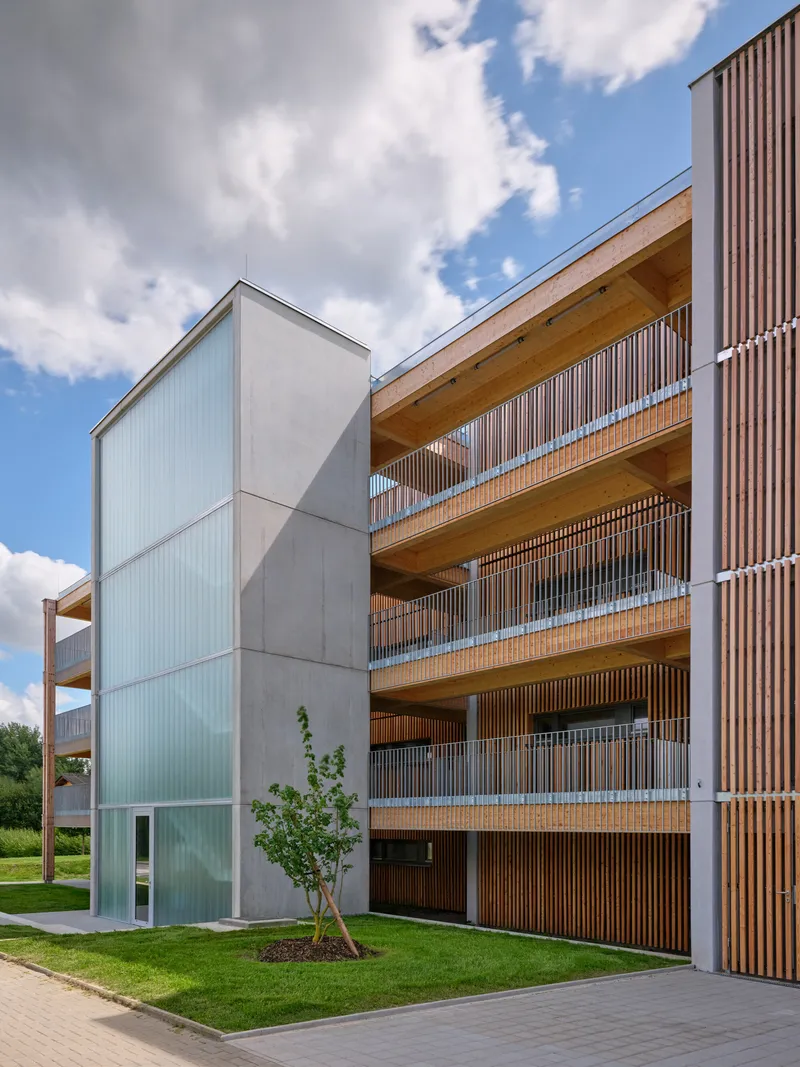 Four-story facade with wooden balconies, vertical slat screens, and young tree in grass lawn