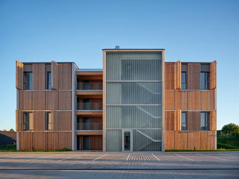 Street elevation showing timber slat volumes flanking a central translucent corrugated stair tower at dusk