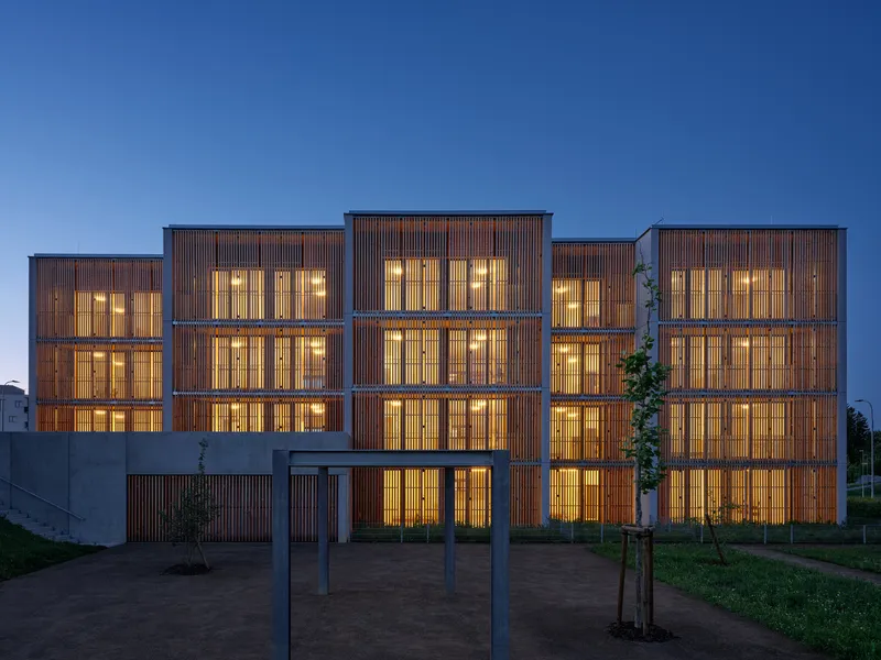 Front facade showing timber louvered screens and interior lighting at dusk with young trees in foreground