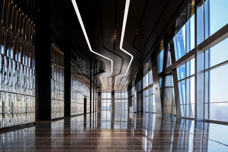 High-rise corridor with floor-to-ceiling glass, decorative metal screens and zigzag illuminated ceiling detail at dusk