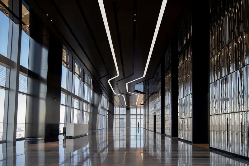 Corridor with floor-to-ceiling glass walls and illuminated metal screens beneath angular ceiling lights