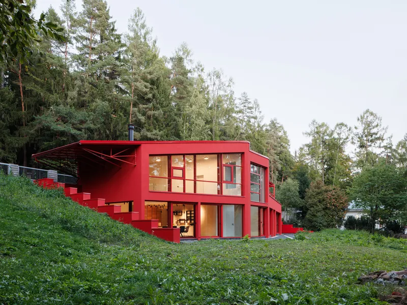 Two-story red facade with glazed openings and exterior staircase set into sloping terrain at dusk