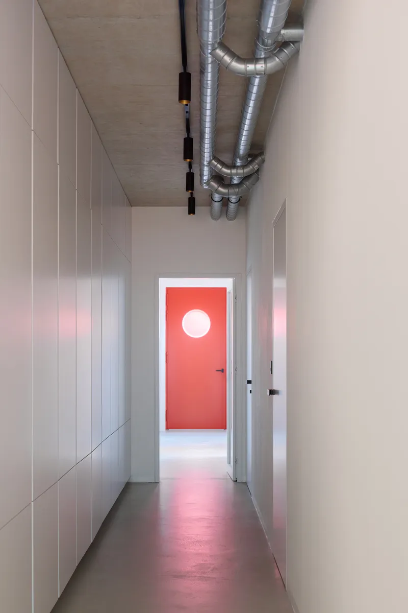 Narrow hallway with exposed ductwork on the ceiling and red door with circular window at end