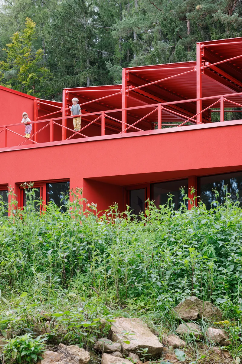 Red steel terrace with cross-braced railing where two children stand among dense forest vegetation