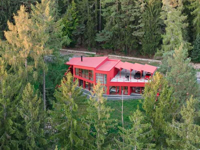 Aerial view of the red metal-clad house with sawtooth roof nestled among conifers in autumn