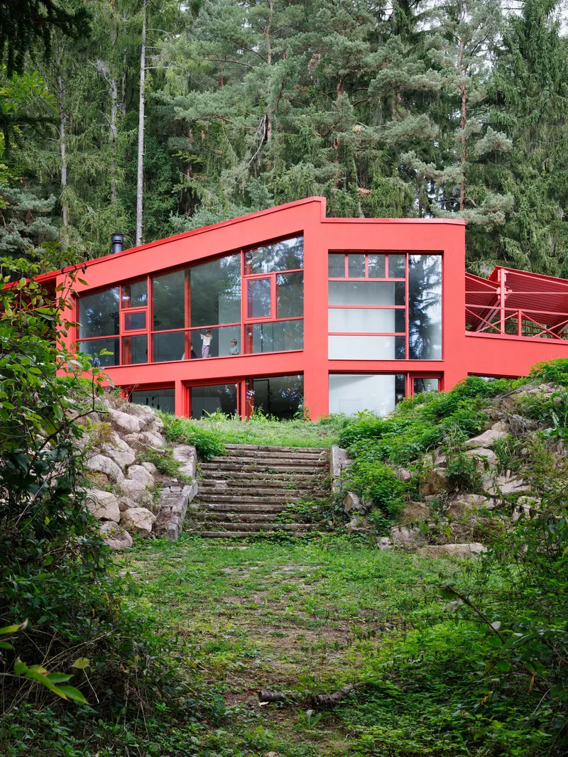 Red steel-framed facade with floor-to-ceiling glazing above a moss-covered stone stairway