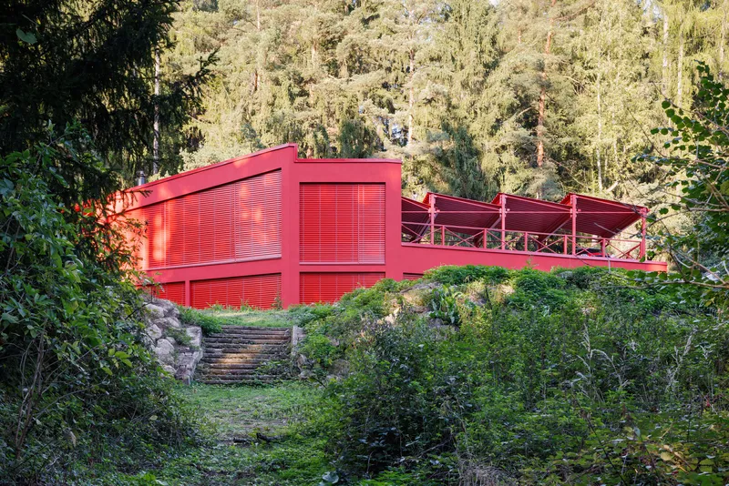 Red painted volume with slatted screens and cantilevered deck nestled into a forested hillside