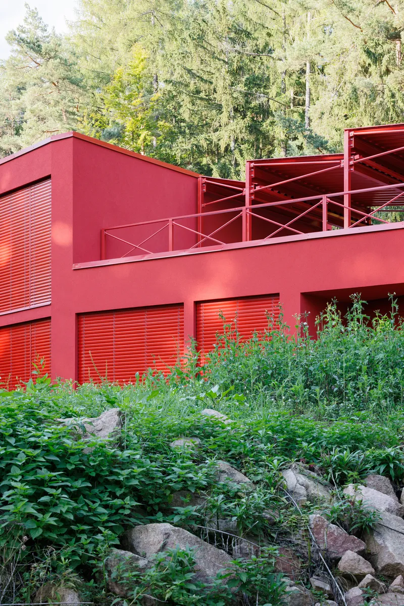 Red metal facade with louvered shutters and upper terrace railing set against rocky planted slope