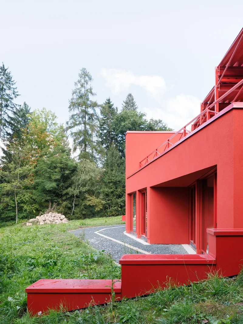 Red painted facade with stepped base and curved gravel driveway through wooded surroundings