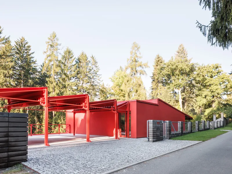 Street view of the red steel carport structure and gabled volume beside cobblestone paving