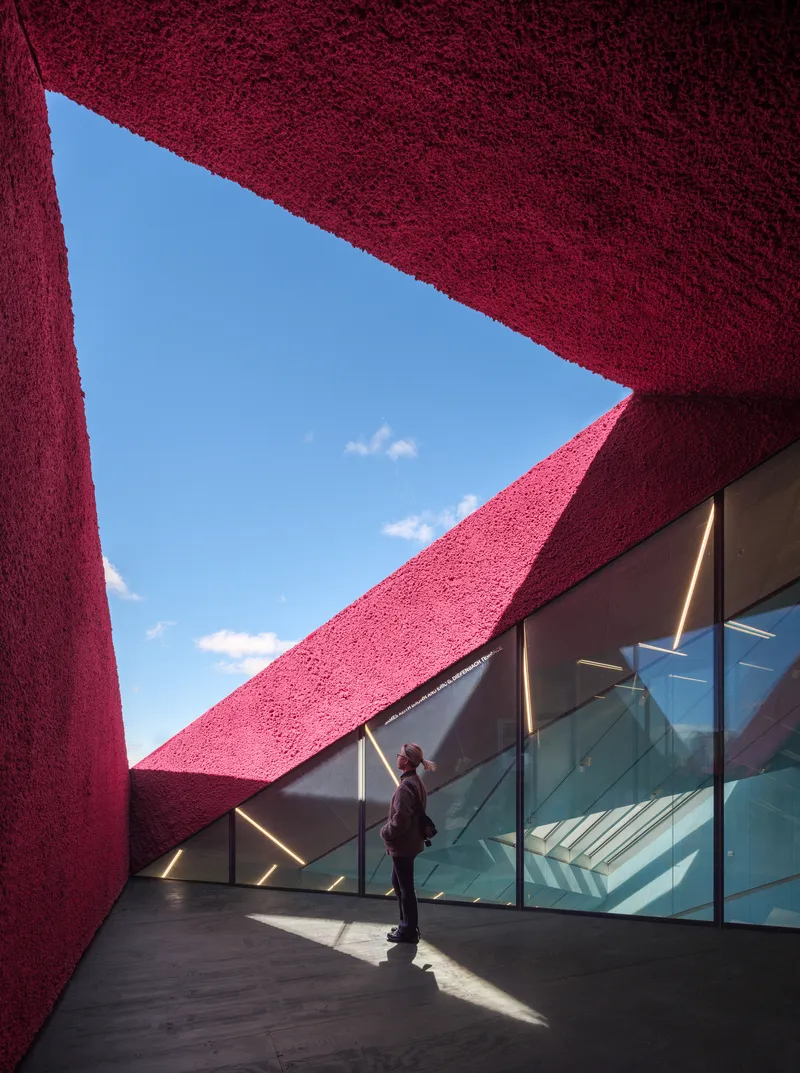 Triangular courtyard framed by textured burgundy walls with a figure standing near glazed facade in sunlight