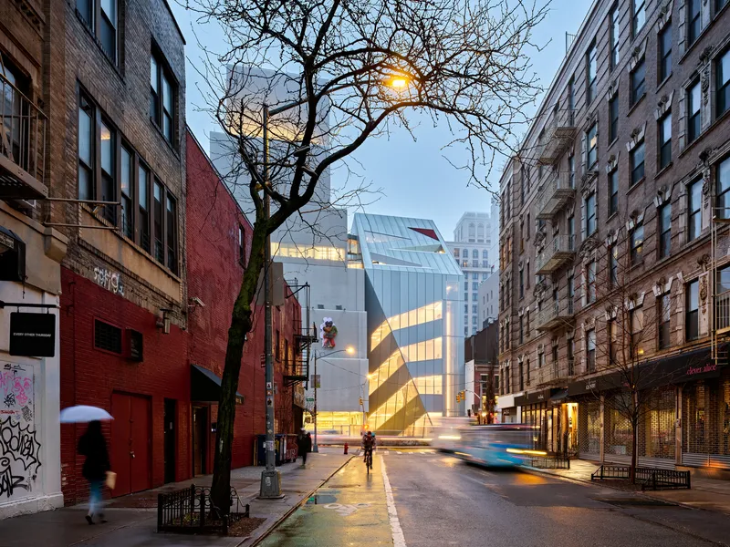 Street view of faceted glass facade at dusk with bare tree and pedestrians between brick buildings