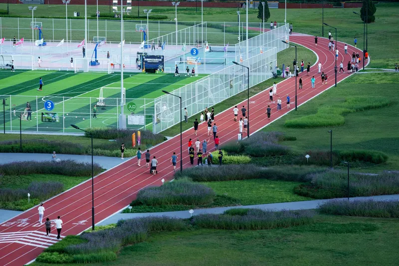 Red running track curving through green lawn with mesh-fenced sports courts and people exercising