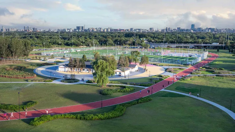 Winding red track alongside curved white path through parkland with distant city skyline under cloudy sky