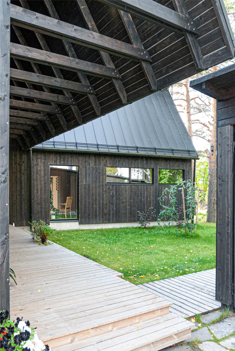 View from covered timber walkway through to charred wood pavilion across lawn courtyard in dappled sunlight