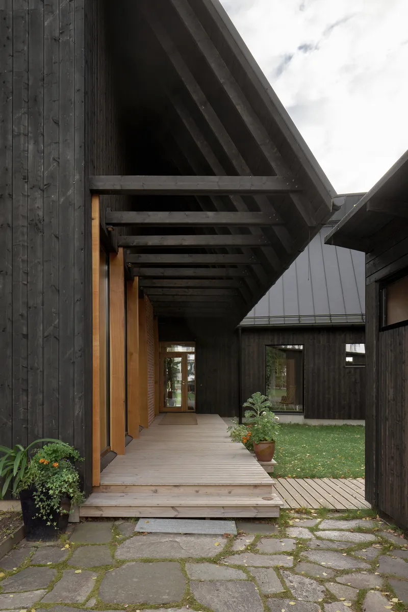 Covered timber walkway with exposed beams leading past planted bed toward charred wood volumes on overcast day