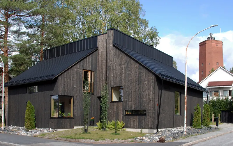 Street facade of black timber-clad volumes with gabled metal roof and scattered window openings