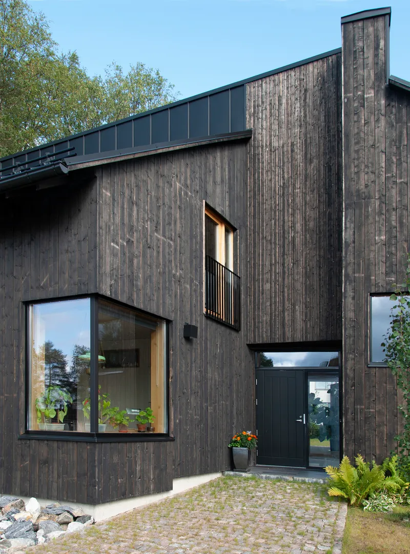 Entrance facade featuring vertical timber boards, projecting window bay with steel railing, and cobblestone paving