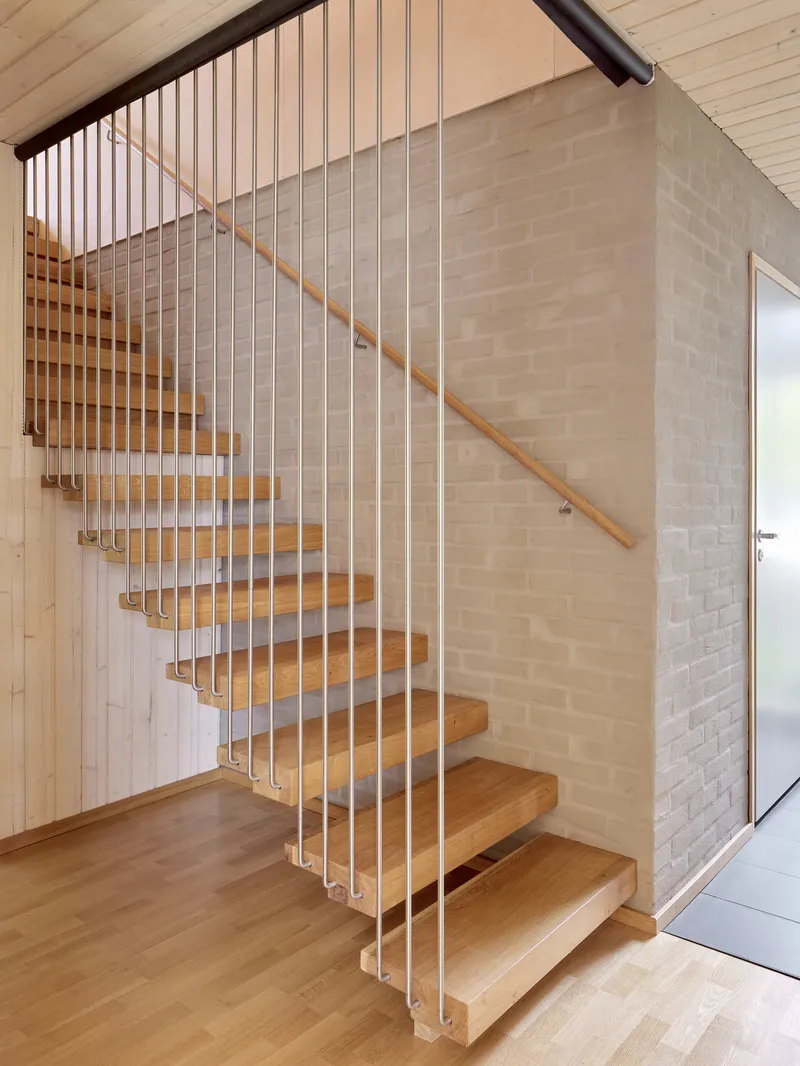 Open-tread timber stair with vertical metal rod balustrade and wooden handrail against pale brick wall