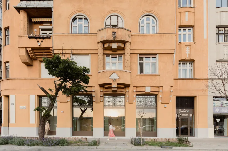 Street facade with peach-toned stucco, arched windows, rounded bay balconies, and a pedestrian passing beneath a tree