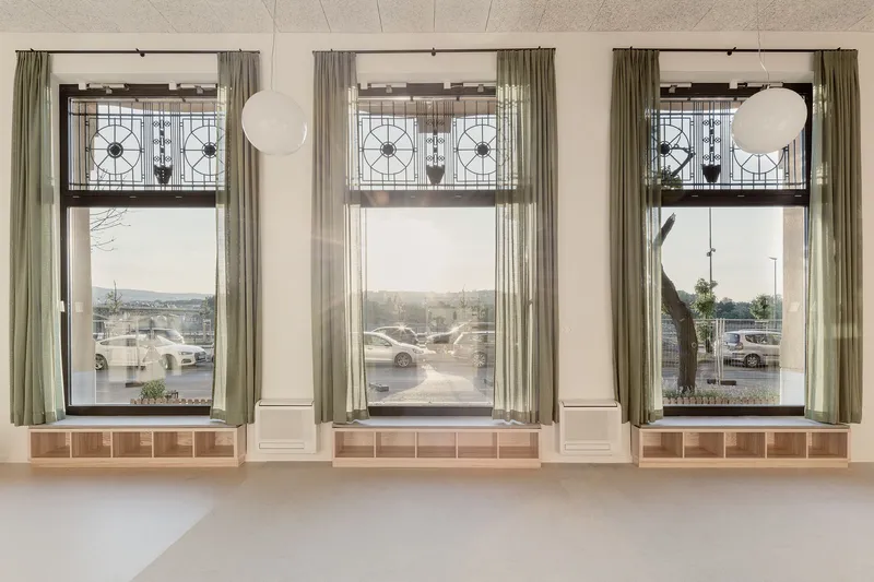 Three tall windows with geometric transom panels and sage curtains overlooking a parking lot in daylight