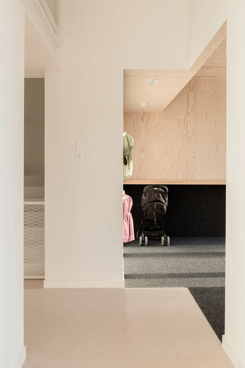 Plywood-clad alcove with integrated bench and dark carpet beside a white plaster corridor with a stroller